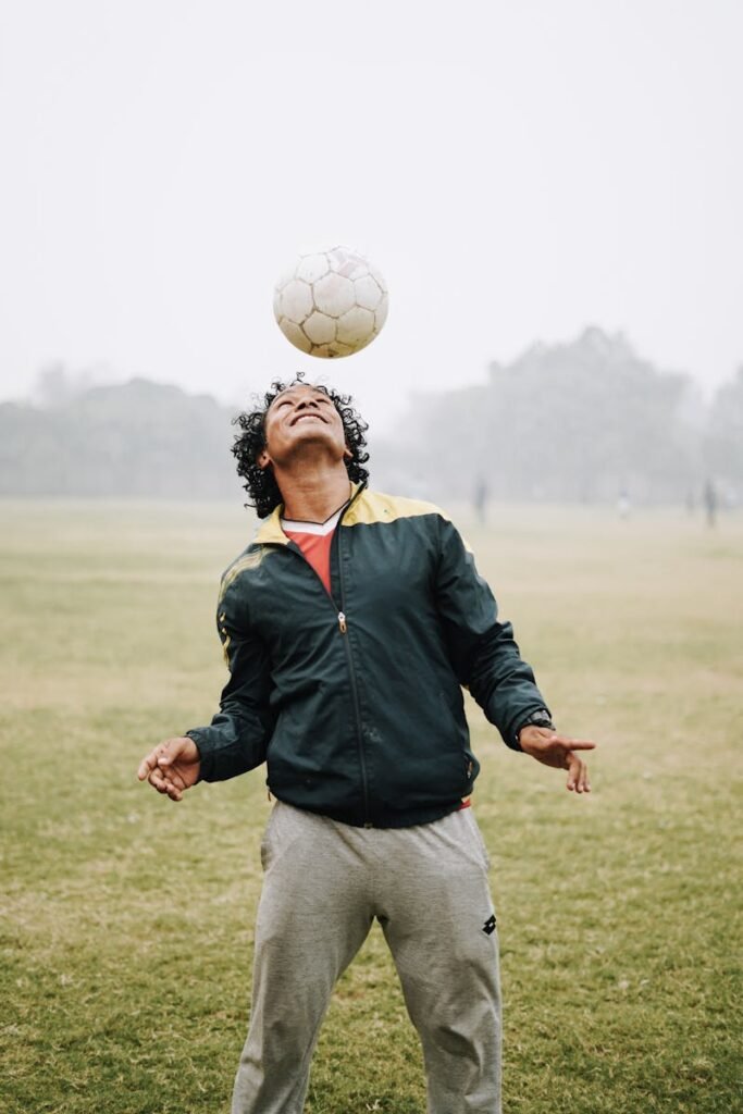 Smiling football male player with curly hair wearing sportswear hitting soccer ball with head while playing on field