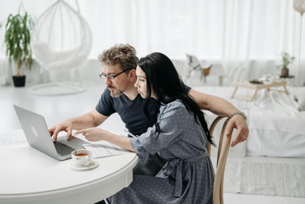 A man and woman sitting indoors, collaborating on a laptop at a table with coffee.